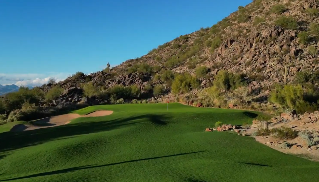 Scenic golf green with sand traps, set against a rocky hillside and blue sky.