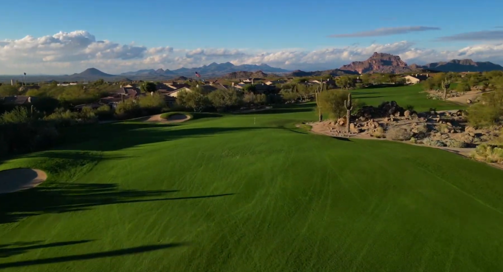 Wide view of a lush green golf fairway surrounded by desert cacti and distant mountains.