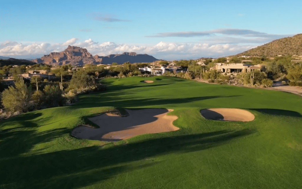 Aerial perspective of a sprawling green golf fairway with large sand traps and mountain backdrop.